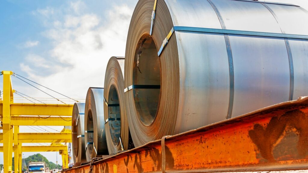 Large rolls of steel are lined up on a rusted orange platform under a clear sky. Yellow industrial structures are visible in the background.