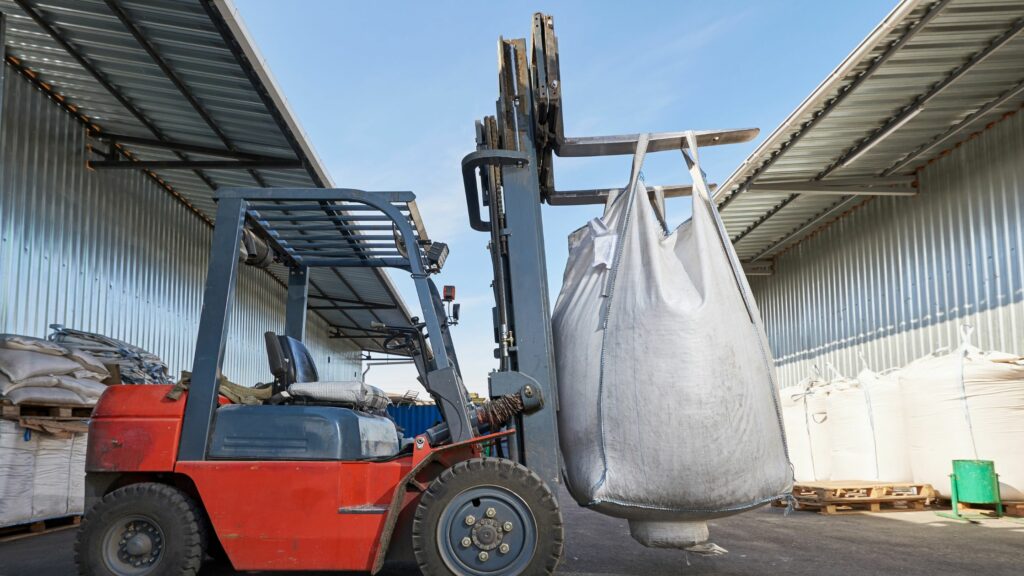 A red forklift is lifting a large white industrial sack in an outdoor warehouse area. The warehouse has a corrugated metal roof and walls, and other sacks and pallets are visible in the background. The sky is clear and blue.