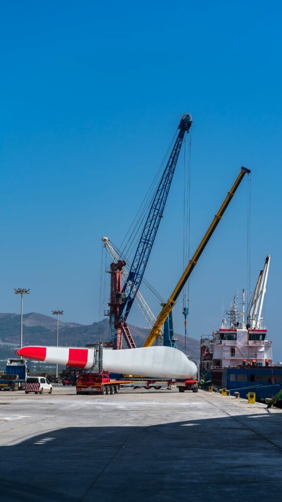 A large wind turbine blade is being transported on a red trailer at an industrial port known for handling break bulk cargo. Several cranes are positioned nearby under a clear blue sky, with a docked ship and hills visible in the background.