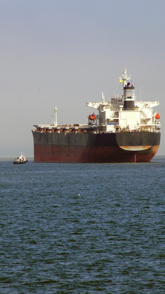 A large cargo ship is anchored in a body of water with a small boat nearby on the left. The sky is clear, and the water appears calm. The cargo ship has visible containers and equipment on its deck.