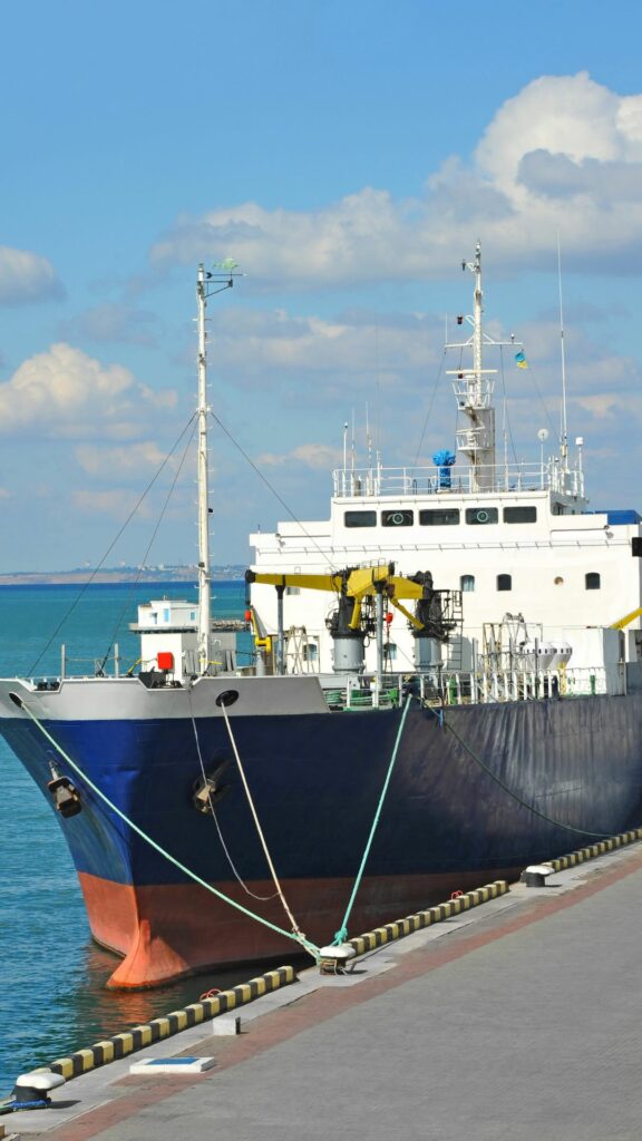 A large cargo ship is docked at a pier on a sunny day. The ship has multiple antennas and masts, and its hull is painted dark blue with a red bottom. The pier has yellow and black safety markings, and the water is calm with a few cloud patches in the sky.