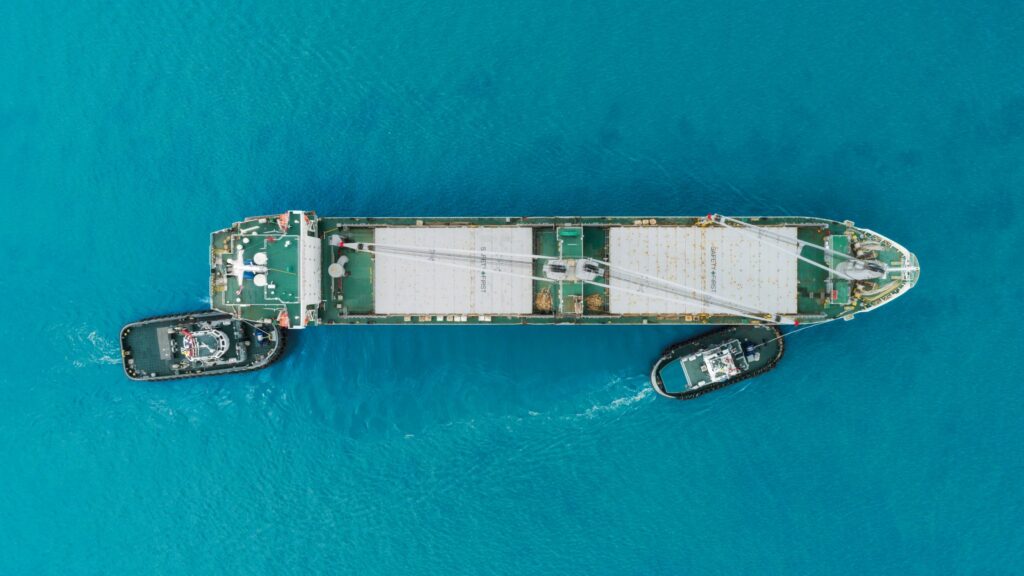 Aerial view of a large cargo ship being guided by two tugboats in clear blue waters. The ship's deck appears to be carrying containers under tarps. The water is vibrant blue, and there are wakes trailing behind the tugboats.