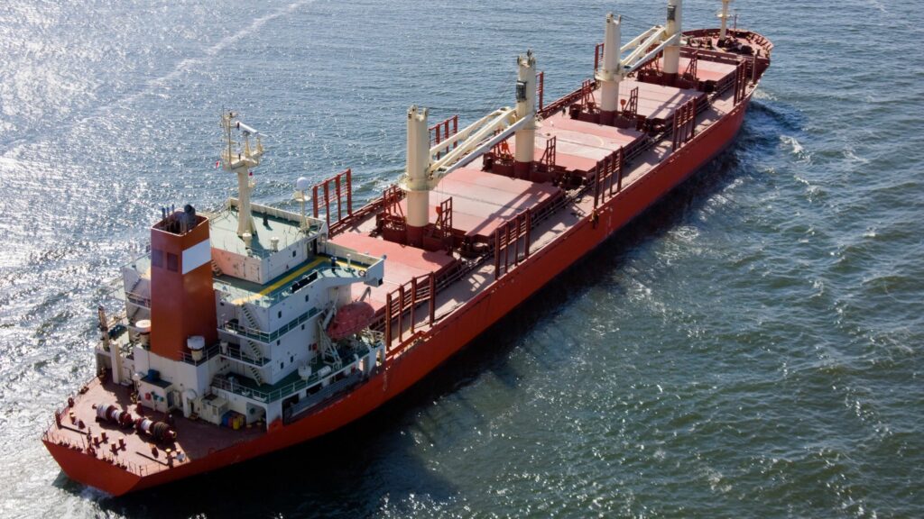 A large cargo ship with red and white coloring sails on a body of water. The ship has multiple compartments and cranes for loading and unloading goods. The surrounding water is calm and sunlit.