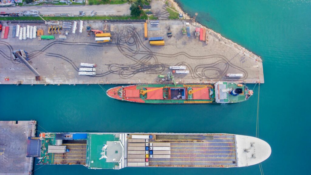 Aerial view of two docked cargo ships at a port. The dock area displays various storage containers and equipment, as well as visible tire marks. The turquoise water contrasts with the grey of the dock and the colorful cargo on the ships.