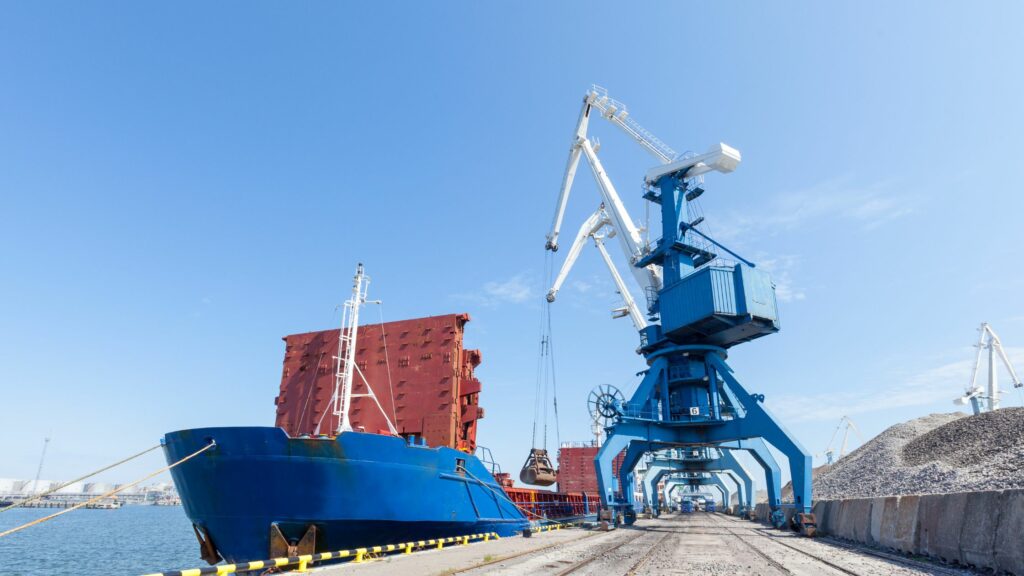 A large blue cargo ship is docked at a port with cranes loading or unloading materials. The surrounding area shows stacked containers and piles of materials, under a clear blue sky.