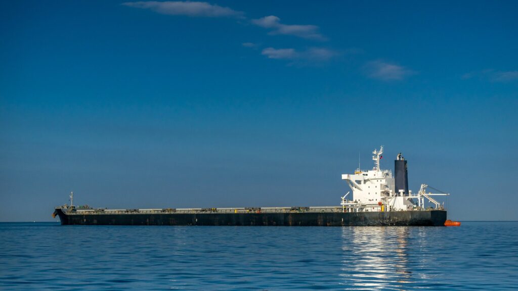 A large cargo ship floats on calm, clear blue water under a clear blue sky with a few clouds. The ship is painted in shades of black and white and has multiple structures on its deck. No other ships or landmasses are visible in the background.