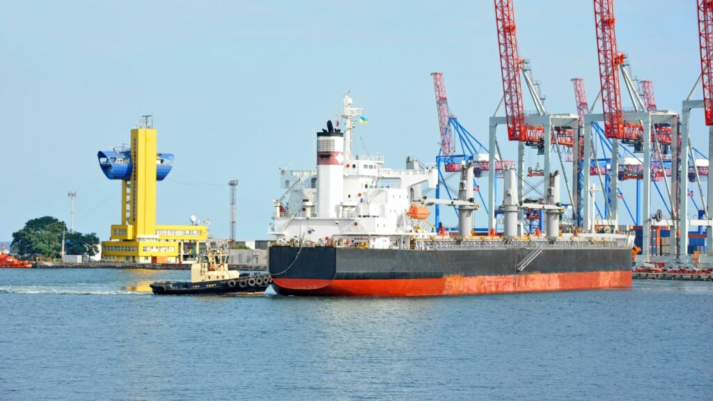 A large cargo ship is docked at a busy port with multiple cranes in the background. There's also a smaller tugboat beside the ship and a yellow building with a blue feature near the water. The weather appears clear with calm water conditions.