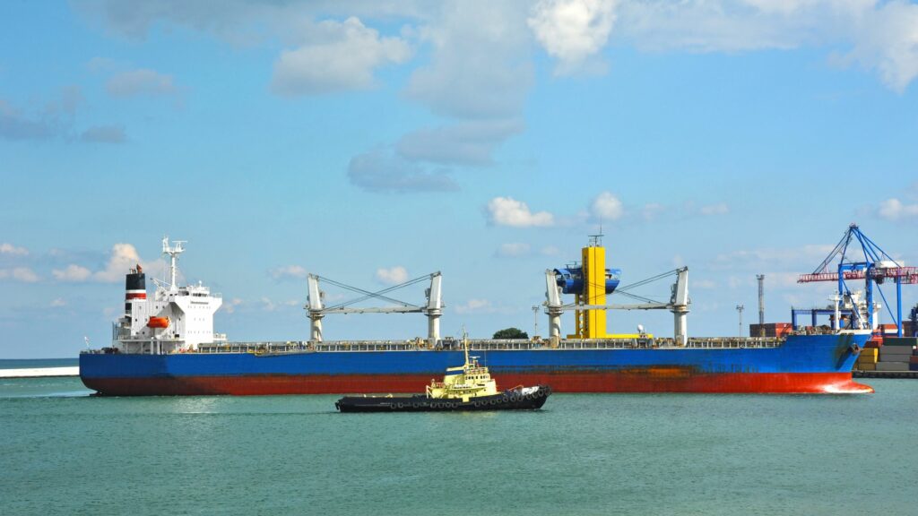 A large cargo ship with a red and blue hull is anchored in a port. A smaller tugboat is in front of it, assisting in navigation. Industrial cranes and port infrastructure are visible in the background under a blue sky with scattered clouds.