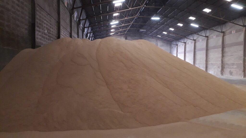 A large pile of sand is stored inside an industrial warehouse with a high ceiling and exposed steel beams. The walls are made of concrete blocks, and several lights are affixed to the ceiling, illuminating the storage area.