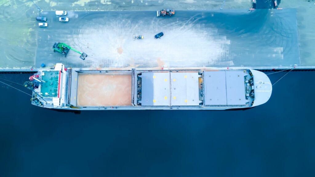 An overhead view of a cargo ship docked at a port. The ship has two large cargo holds with open covers, and there are several vehicles and pieces of equipment on the dock nearby. The water surrounding the dock and the ship is a deep blue color.