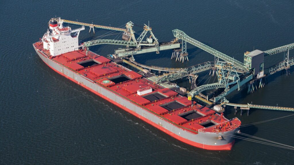 An aerial view of a large red cargo ship docked at a loading facility in the water. The ship has multiple cargo holds and is being loaded or unloaded by several green cranes. The surrounding water is calm and there are no other vessels nearby.