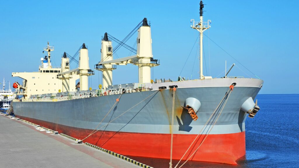 A large cargo ship is docked at a port. The ship has a gray hull with a red bottom, and several cranes and masts on its deck. Ropes secure the vessel to the concrete pier. The scene is backed by a clear blue sky and calm sea.