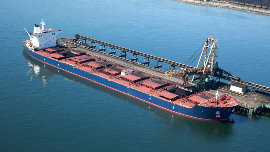 A large cargo ship is docked at a port with an industrial loading structure extending over its deck. The ship is blue with a red bottom and has several compartments on its deck. The calm water reflects the vessel and surrounding structures.