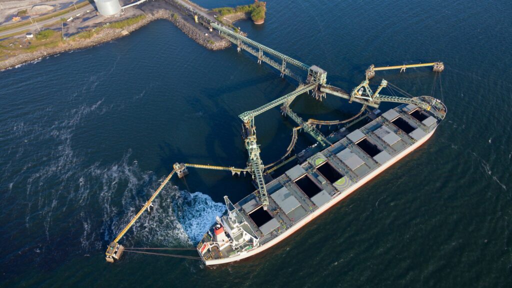 Aerial view of a large cargo ship docked at an industrial port. The ship is being loaded or unloaded via multiple conveyor belts extending from the port's infrastructure. The surrounding water and shoreline are visible, with some land structures in the background.