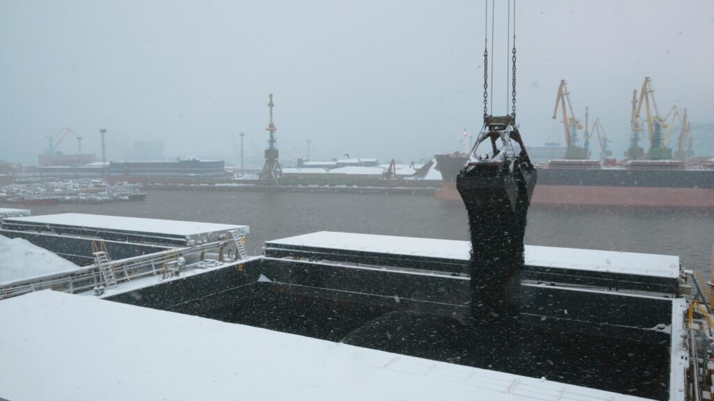 A large mechanical claw is depositing a load of coal into a cargo hold of a ship during snowfall at a port. Cranes and other industrial infrastructure are visible in the background, partially obscured by falling snow.