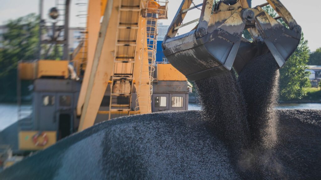 An industrial claw machine unloads black granular material from a large container on a construction site. A yellow crane supports the activity in the background, with trees and a body of water visible in the distance.