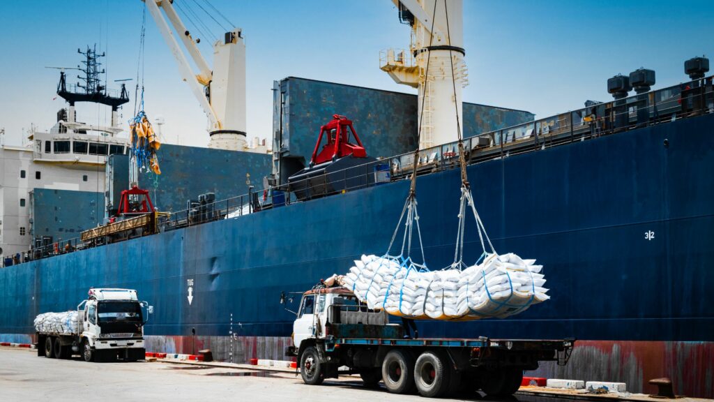 Two trucks are being loaded with large bags of goods using a crane from a cargo ship. The scene takes place at a commercial port under a clear sky, showcasing a logistics operation involving maritime transport and ground vehicles.