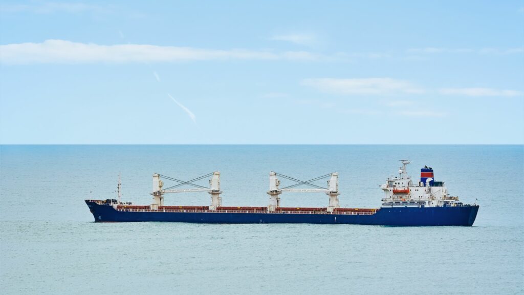 A large cargo ship with blue hull and white superstructure is sailing on a calm sea. The sky is clear with a few clouds. The ship has two cranes positioned midship along its deck.