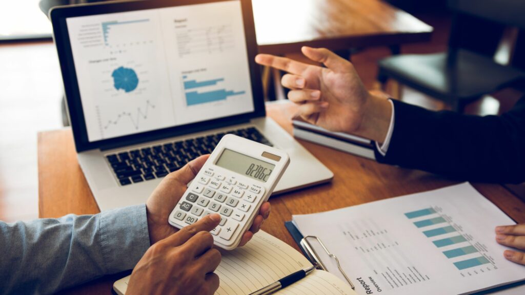 Two people are discussing financial data in an office. One person is holding and using a calculator, while the other points at a laptop screen displaying charts. A notebook and financial report with charts are lying on the table.