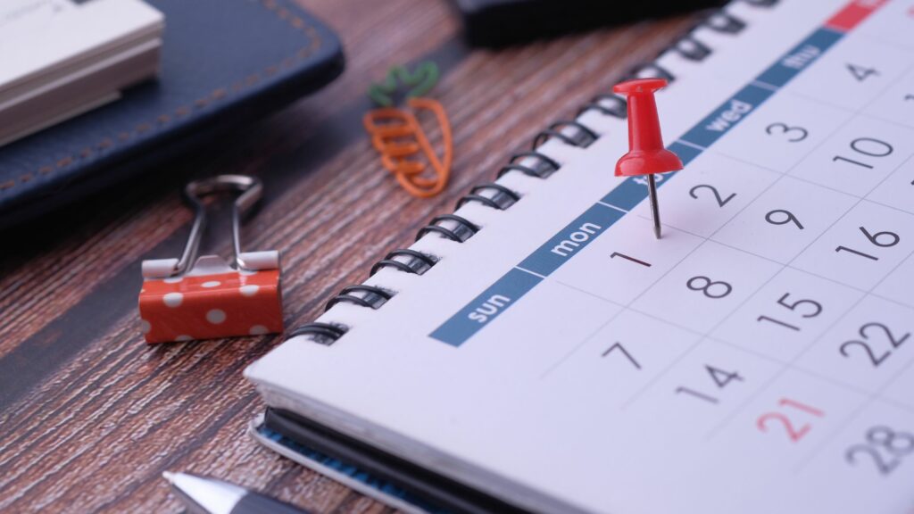 A close-up of a spiral-bound calendar open to a month with dates visible. A red pushpin marks Monday, the 1st. Nearby are a polka-dotted binder clip, a carrot-shaped paperclip, and a pen resting on top of a leather notepad—tools essential for planning and logistics—on a wooden surface.