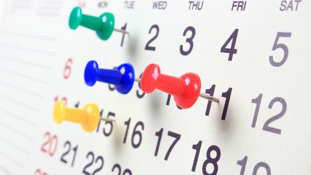 A close-up of a wall calendar with push pins on specific days. The pins, essential for coordinating logistics, are colored yellow, blue, green, and red, pinned on various dates. The dates and days of the week laycan are clearly visible in black text on a white background.