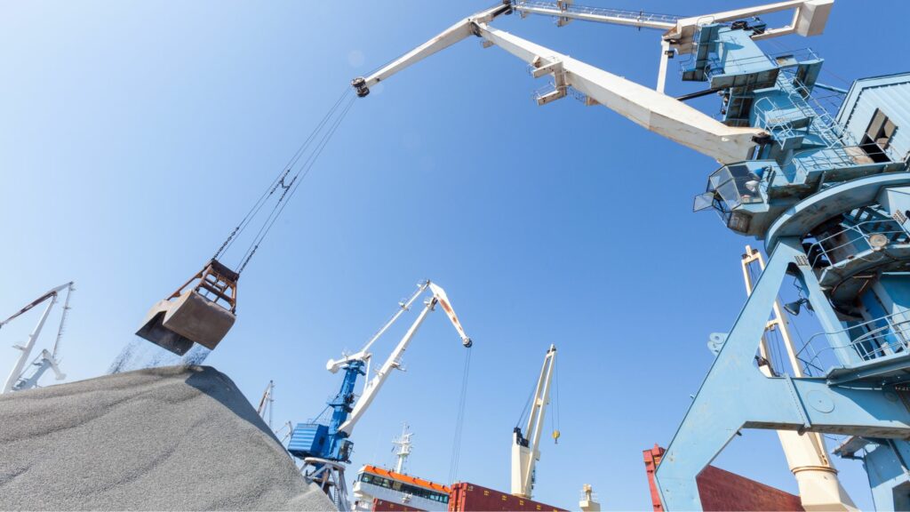 A large, industrial crane is operating at a shipping port, lifting a load of material. Other cranes and cargo containers are visible in the background under a clear blue sky.