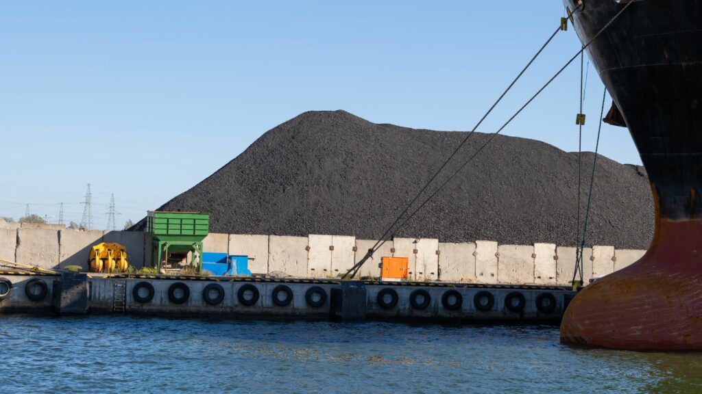 A large pile of coal sits behind a concrete barrier at a port. Part of a ship is visible on the right, with mooring lines extending from it. The water in the foreground is calm, and various industrial containers and equipment are positioned near the barrier.