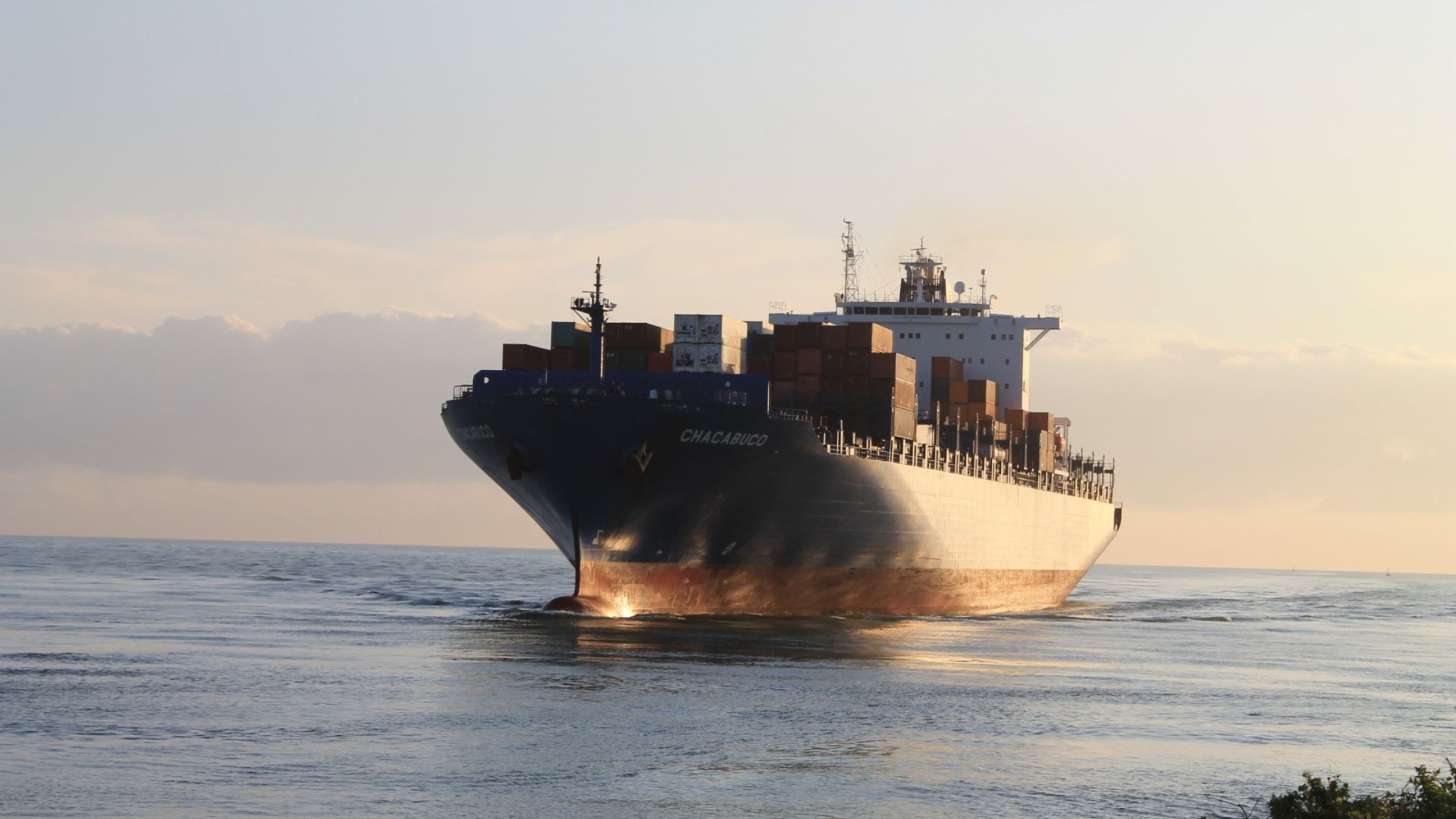 A large cargo ship, named "CHACABUCO," sails on the water during sunset. The ship is loaded with shipping containers and navigates forward, creating small waves in its wake. The sky in the background has a mixture of light clouds and warm light from the setting sun.