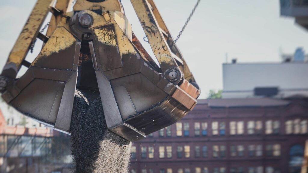 A close-up of an industrial claw crane releasing gravel or small rocks. The background shows out-of-focus buildings, with one structure partially visible. The scene appears to be taking place in an urban construction or industrial area during daytime.