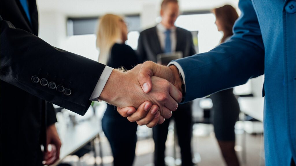 Two people are shaking hands in the foreground of an office setting, perhaps finalizing logistics for a project. Both individuals are dressed in formal business attire. In the blurred background, three other people are engaged in conversation, suggesting a business meeting or event.