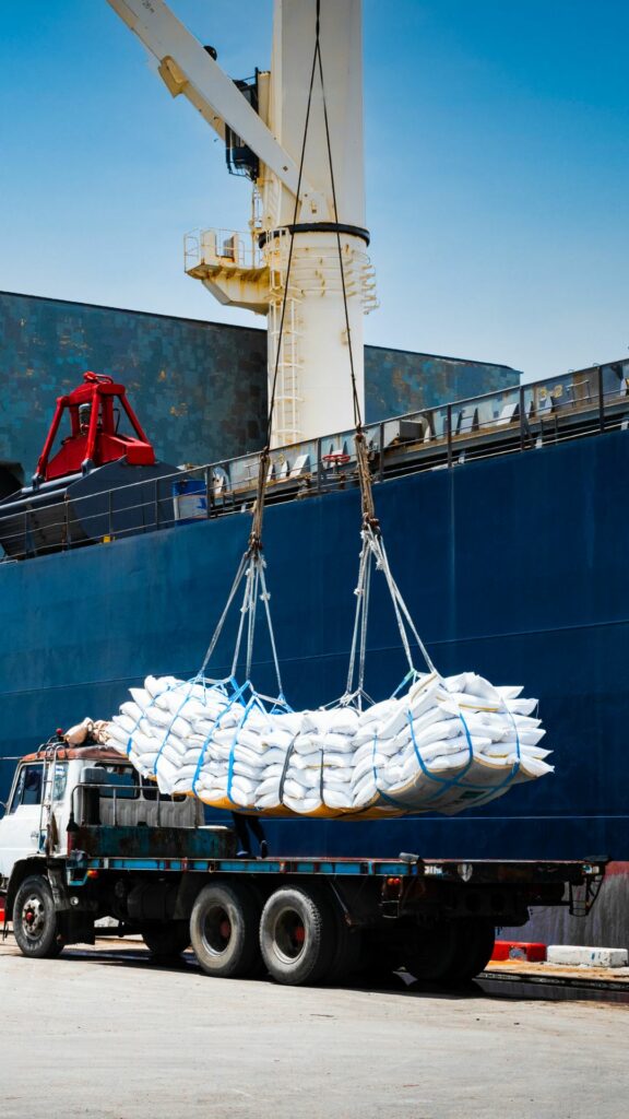 A crane is lifting a large net filled with sacks from the back of a truck at a shipping port. In the background, there is a large cargo ship. The scene indicates the loading or unloading process of goods.