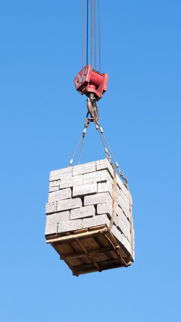 A crane lifting a pallet of grey cinder blocks against a clear blue sky. The pallet is secured with ropes and appears to be in motion.