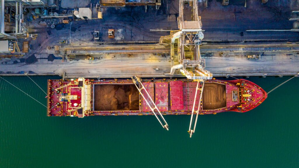 Aerial view of a large cargo ship docked at an industrial port. The ship has open cargo holds filled with material, and cranes extend over it for loading or unloading. The port equipment and infrastructure are visible around the ship.