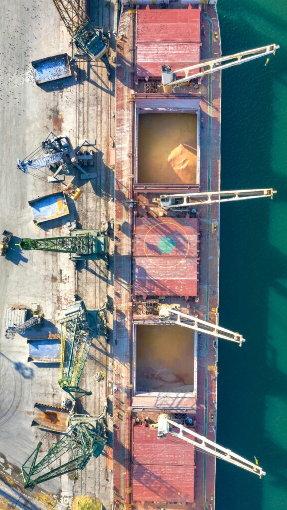 Aerial view of a cargo ship docked at a pier with cranes unloading material from its holds. The ship's deck has three large open compartments filled with yellowish material, possibly grain or sand. The surrounding area includes additional cargo handling equipment.