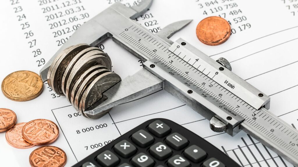 A caliper measuring a stack of coins rests on a financial document with numbers and graphs. Nearby, there are additional coins scattered and a black calculator.