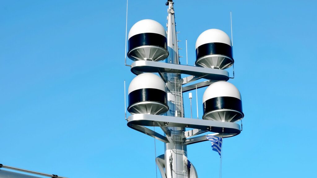 The image shows a yacht mast equipped with four white spherical radar domes mounted in pairs on opposite sides. A Greek flag, symbolizing the vessel's adherence to Admiralty Law, is attached to the structure, flying against a clear blue sky.