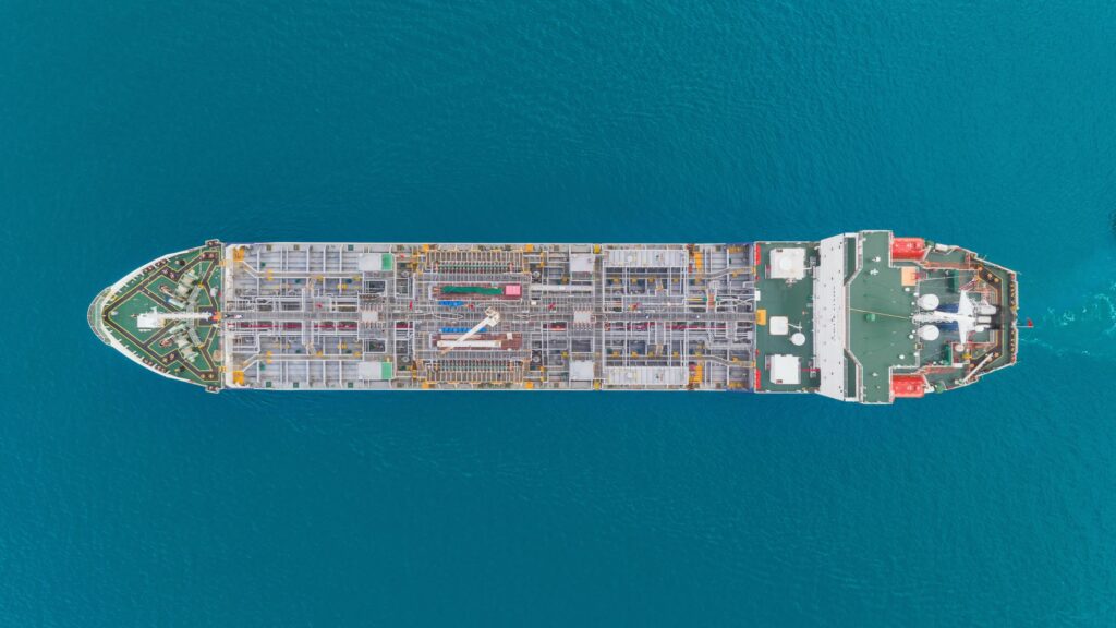 Aerial view of a cargo ship navigating through clear blue waters. The ship has a variety of containers and equipment on deck, and there are visible pathways and structures on the ship's surface. The water surrounding the ship is a deep blue color.