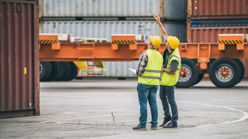 Two workers wearing reflective vests and hard hats stand in an outdoor shipping yard. One worker holds a clipboard while the other points upwards. Shipping containers and a large orange transport vehicle are visible in the background.