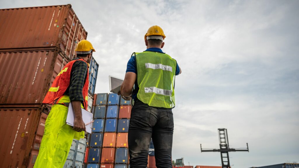 Two workers wearing hard hats and safety vests are standing in front of a stack of shipping containers. One is holding a clipboard and the other a laptop. There is an industrial crane in the background against a cloudy sky.