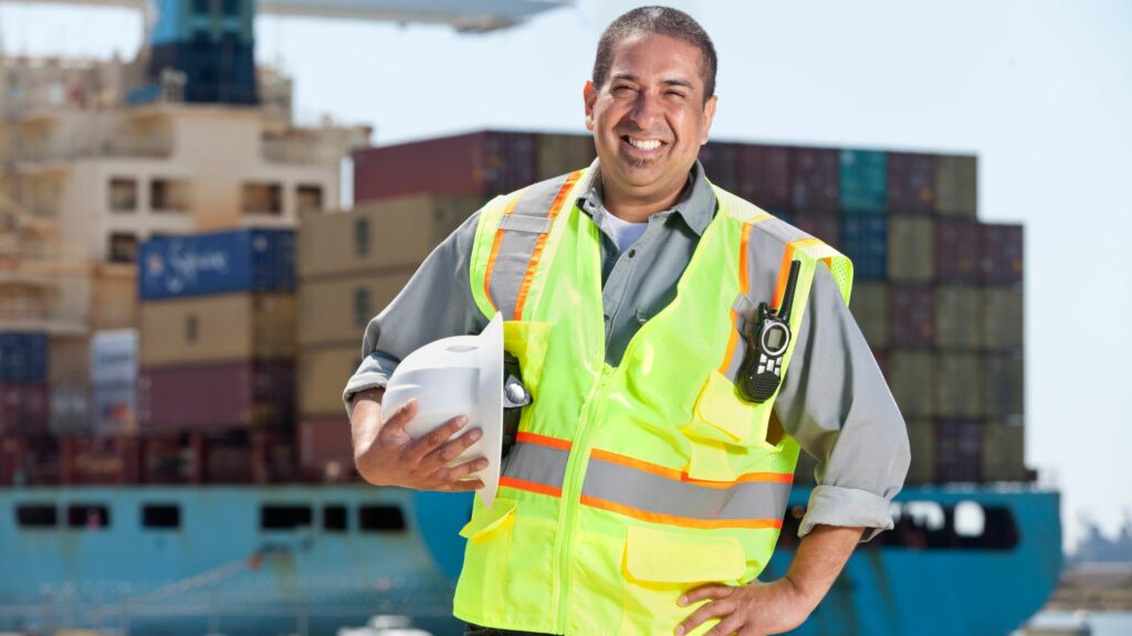 A man wearing a high-visibility safety vest and holding a white hard hat stands in front of a large cargo ship loaded with shipping containers. He is smiling and has a handheld radio clipped to his vest. The setting is likely a dock or shipping yard.