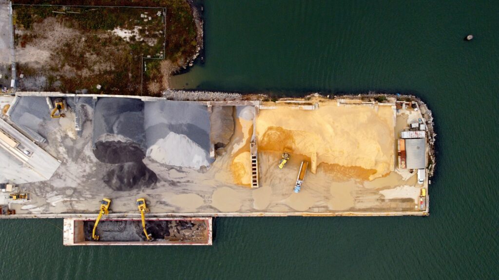 Aerial view of an industrial pier with piles of different colored materials. Several yellow construction vehicles, including excavators, are positioned around the site. The pier is surrounded by water on three sides.