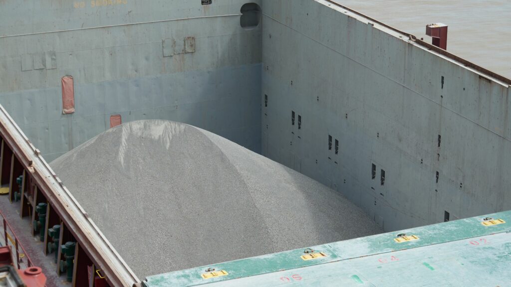 A large cargo ship's hold is partially filled with a mound of grey gravel. The walls of the cargo hold are grey, and some of the hatches on the deck are visible in the foreground. The scene is industrial, suggesting the transportation of bulk materials.