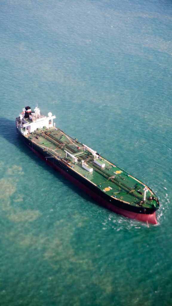 Aerial view of a large cargo tanker ship navigating through a greenish-blue sea. The ship has a dark hull with a white structure on top, and various equipment and containers are visible on the deck. The water is relatively calm around the vessel.