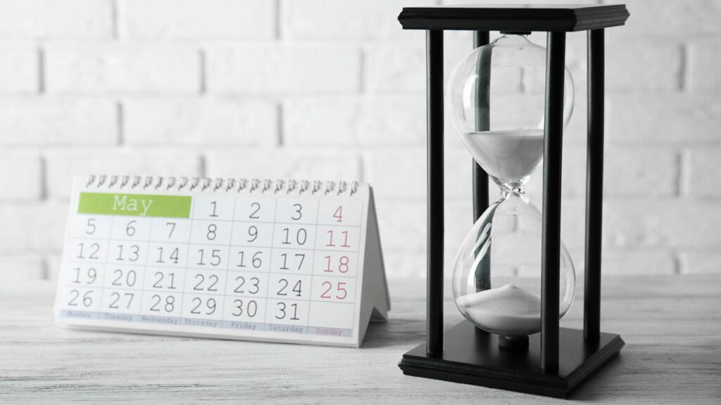 A black hourglass with white sand is placed on a table next to a spiral-bound desk calendar showing the month of May. The background features a white brick wall.