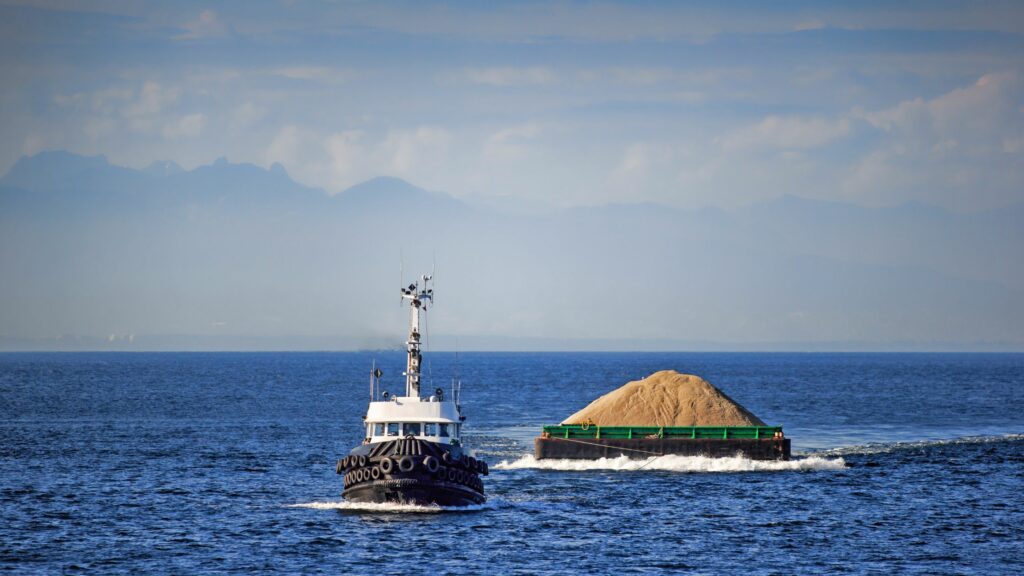 A tugboat pulls a barge loaded with sand across a calm blue sea. The barge has green railings, and the sea appears vast and deep. In the background, there are distant mountains under a partly cloudy sky.