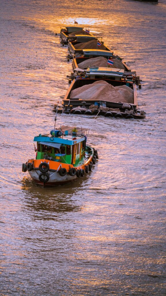 A boat is towing multiple barges loaded with sand along a river at sunset. The water reflects the warm colors of the setting sun, creating a golden hue. The scene captures a working vessel in a peaceful, picturesque setting.