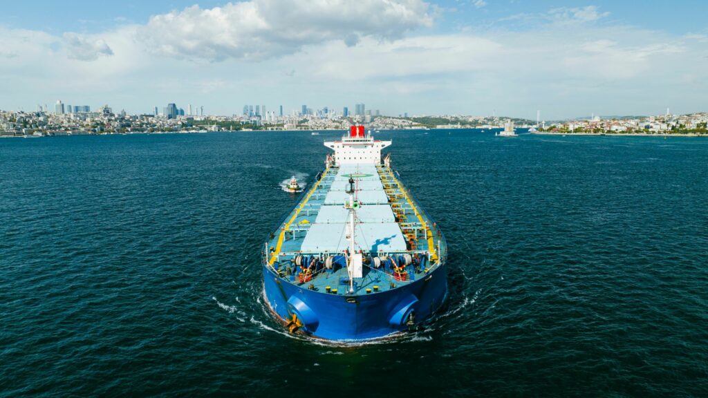 A large blue cargo ship is sailing through a wide body of water towards the horizon. The ship is accompanied by a smaller boat on its left side. A cityscape with numerous buildings and skyscrapers appears in the background under a partly cloudy sky.