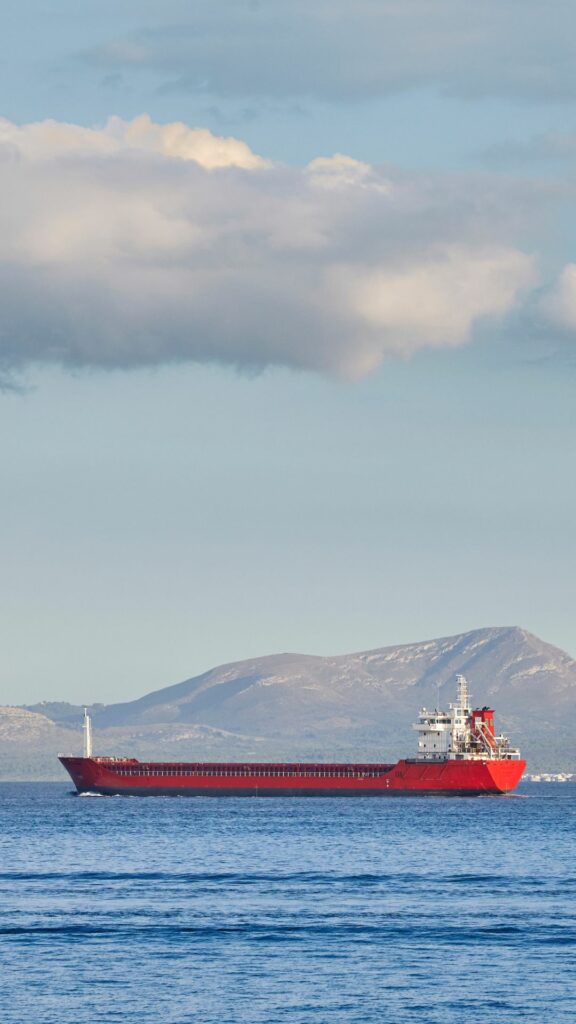 A red cargo ship sails on calm blue waters, with mountainous terrain visible in the distance under a partly cloudy sky.