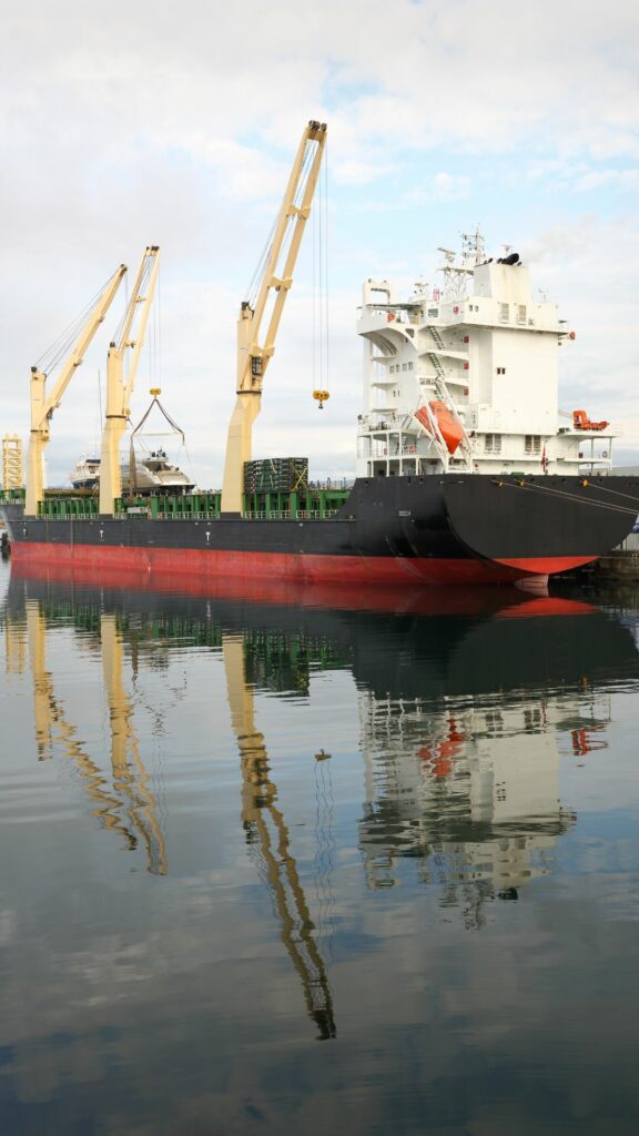 A large cargo ship with two yellow cranes is docked at a port. The ship's hull is black and red, and its reflection is visible in the calm water. The sky is partly cloudy.