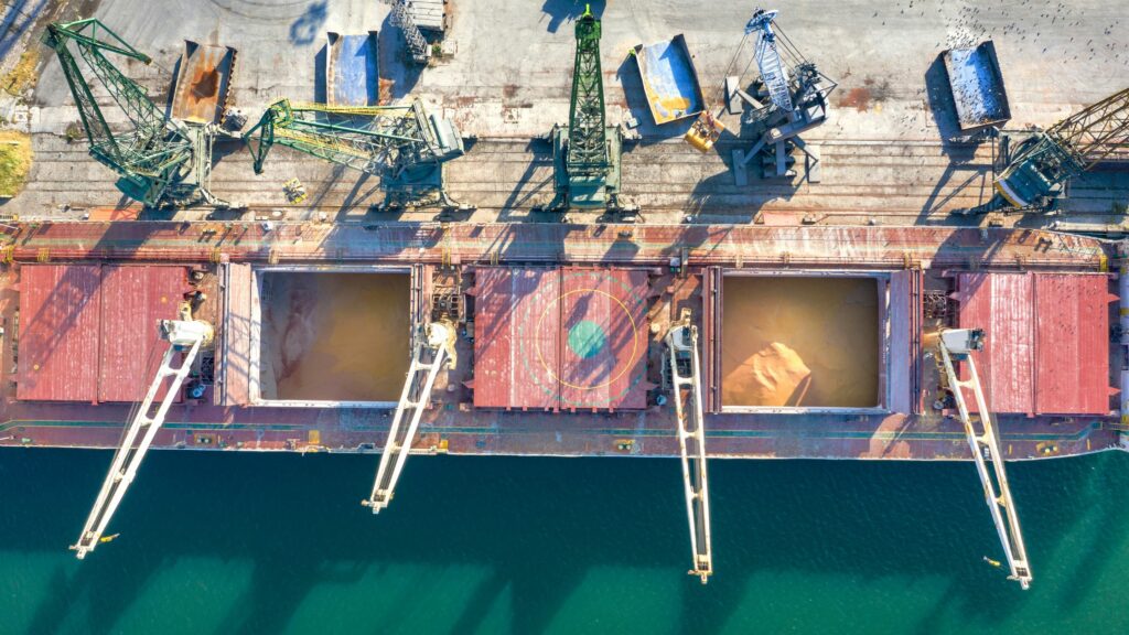 Aerial view of a cargo ship docked at a port with its hatches open. Large cranes are positioned over the holds, likely for loading or unloading goods. The ship is alongside a concrete dock visible in the upper part of the image, adjacent to the water.